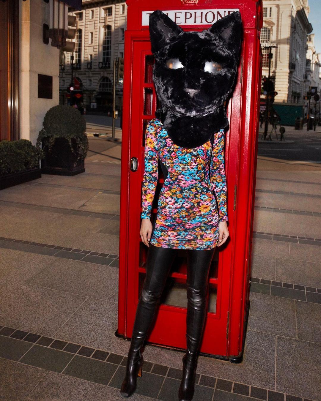 Person in a cat headpiece and floral dress poses next to a red telephone booth on an urban street, creating a surreal fashion moment.
