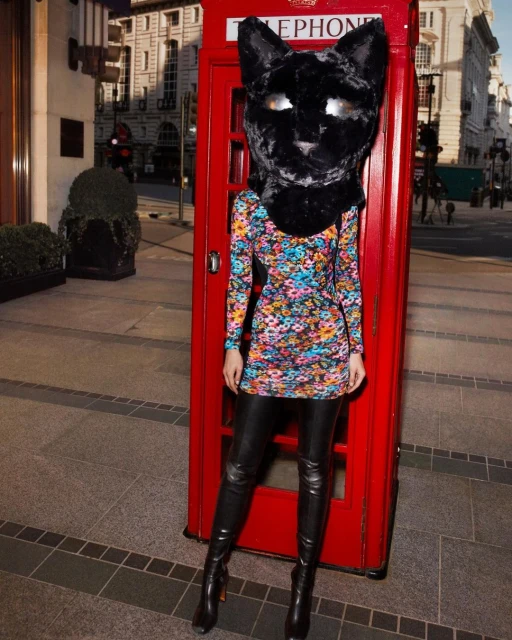 Person in a cat headpiece and floral dress poses next to a red telephone booth on an urban street, creating a surreal fashion moment.