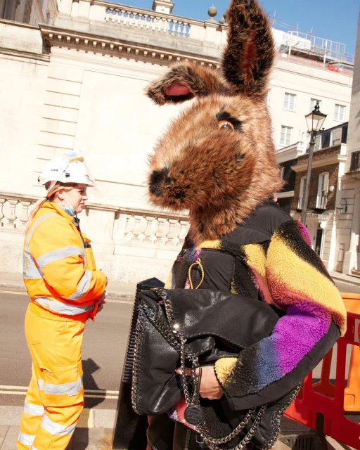 Person in a large brown furry rabbit head stands near a construction worker in an orange jumpsuit on a city street.