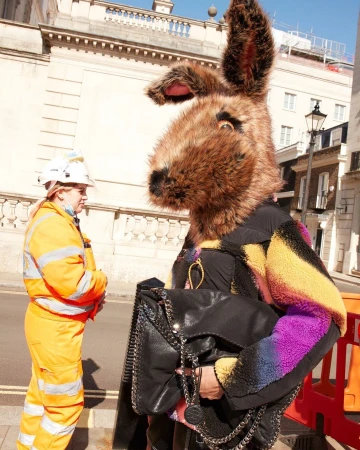 Person in a large brown furry rabbit head stands near a construction worker in an orange jumpsuit on a city street.