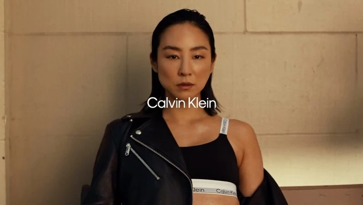 Asian female model in Calvin Klein underwear and leather jacket poses against a neutral wall, showcasing beauty and fashion.