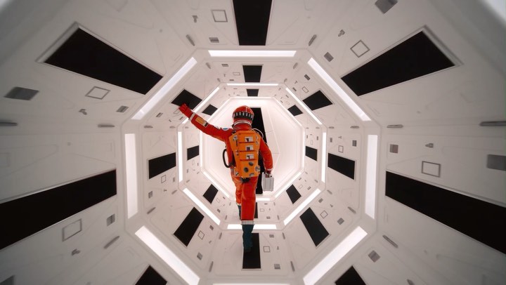 A person in an orange spacesuit waves in a white octagonal tunnel with black rectangular panels in a low-angle shot. Space travel theme.