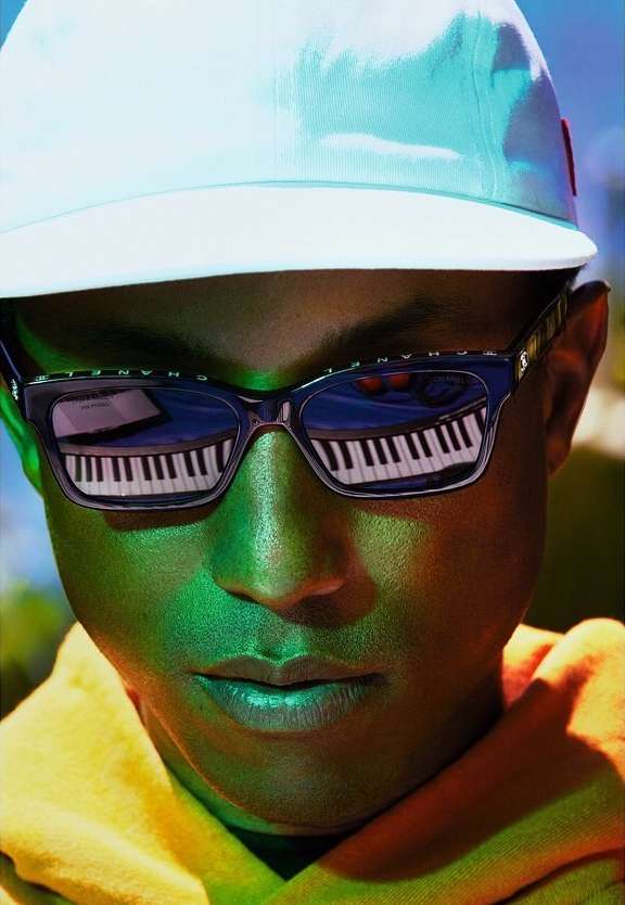 Close-up of an adult wearing sunglasses reflecting a keyboard, a white baseball cap, and a yellow jacket, with green and blue lighting.