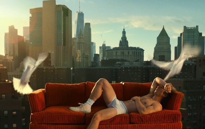 Adult male reclines on orange couch with white doves flying in an urban setting with tall buildings and a blue sky in the daytime.