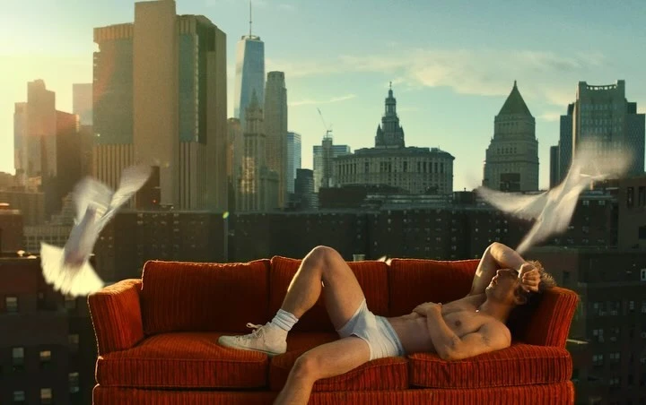 Adult male reclines on orange couch with white doves flying in an urban setting with tall buildings and a blue sky in the daytime.