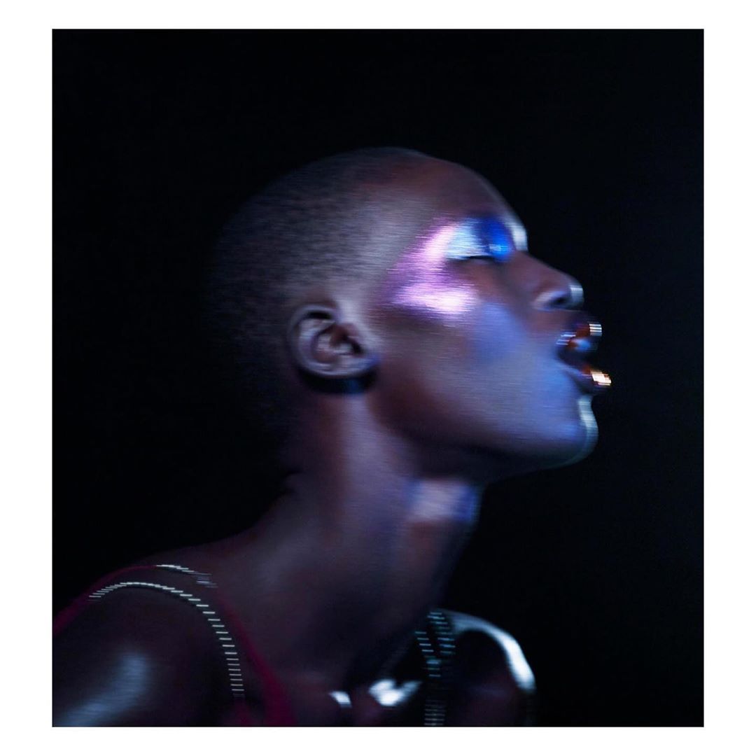 Portrait of a stylish woman with an updo, wearing iridescent makeup and a metal necklace, looking up in a dark, indoor setting.