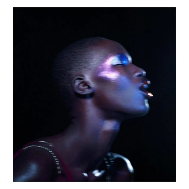 Portrait of a stylish woman with an updo, wearing iridescent makeup and a metal necklace, looking up in a dark, indoor setting.