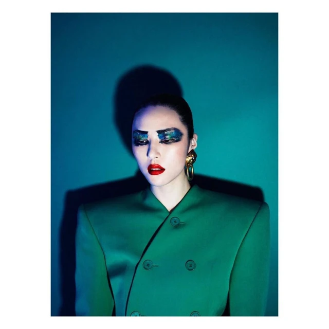 Eye-level studio portrait of a young woman with dramatic makeup, gold earrings, and green jacket, against a teal backdrop with shadow.