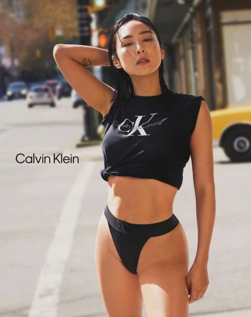 Asian woman poses in Calvin Klein top and underwear on city street with cars and yellow taxi cab in background during daytime.