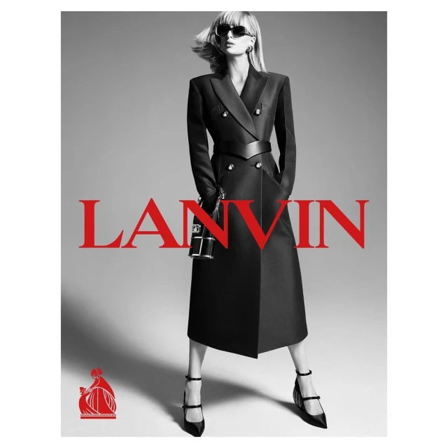 Fashion model posing in a long black coat with a wide belt, black heels, and sunglasses in a studio fashion advertisement.