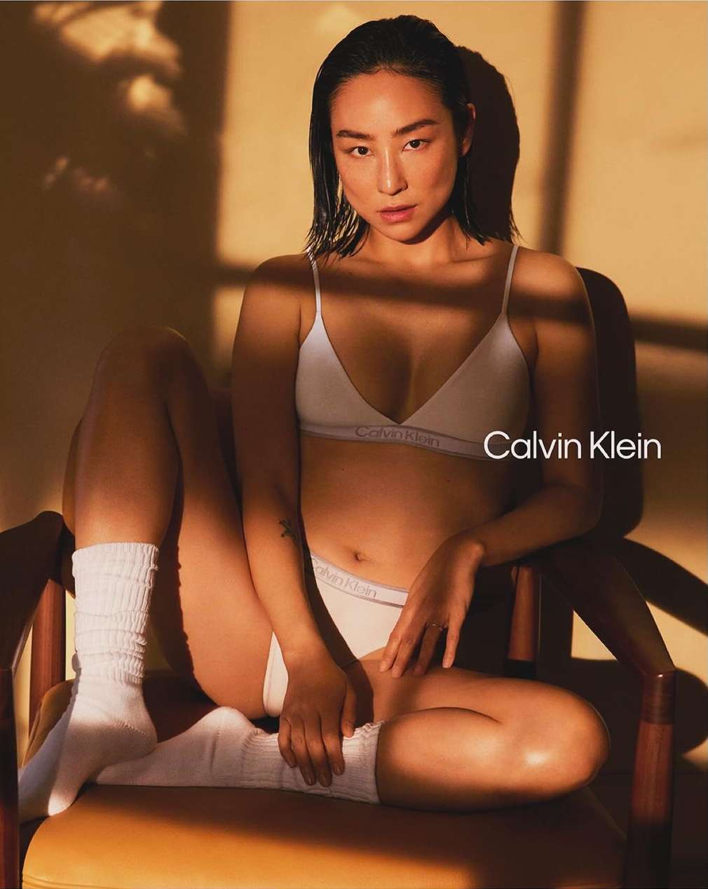 A woman in white Calvin Klein underwear sits on a chair in sunlight. The intimate apparel campaign features a modern aesthetic.