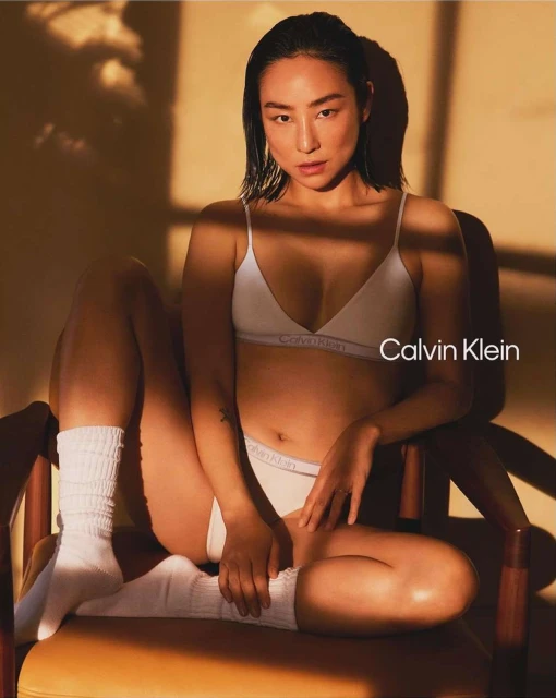 A woman in white Calvin Klein underwear sits on a chair in sunlight. The intimate apparel campaign features a modern aesthetic.