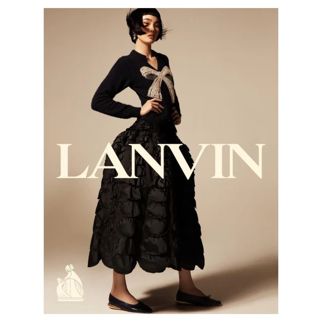 Fashion editorial shot of an adult woman in a black dress with a large bow on her sweater, posing indoors in a studio environment.