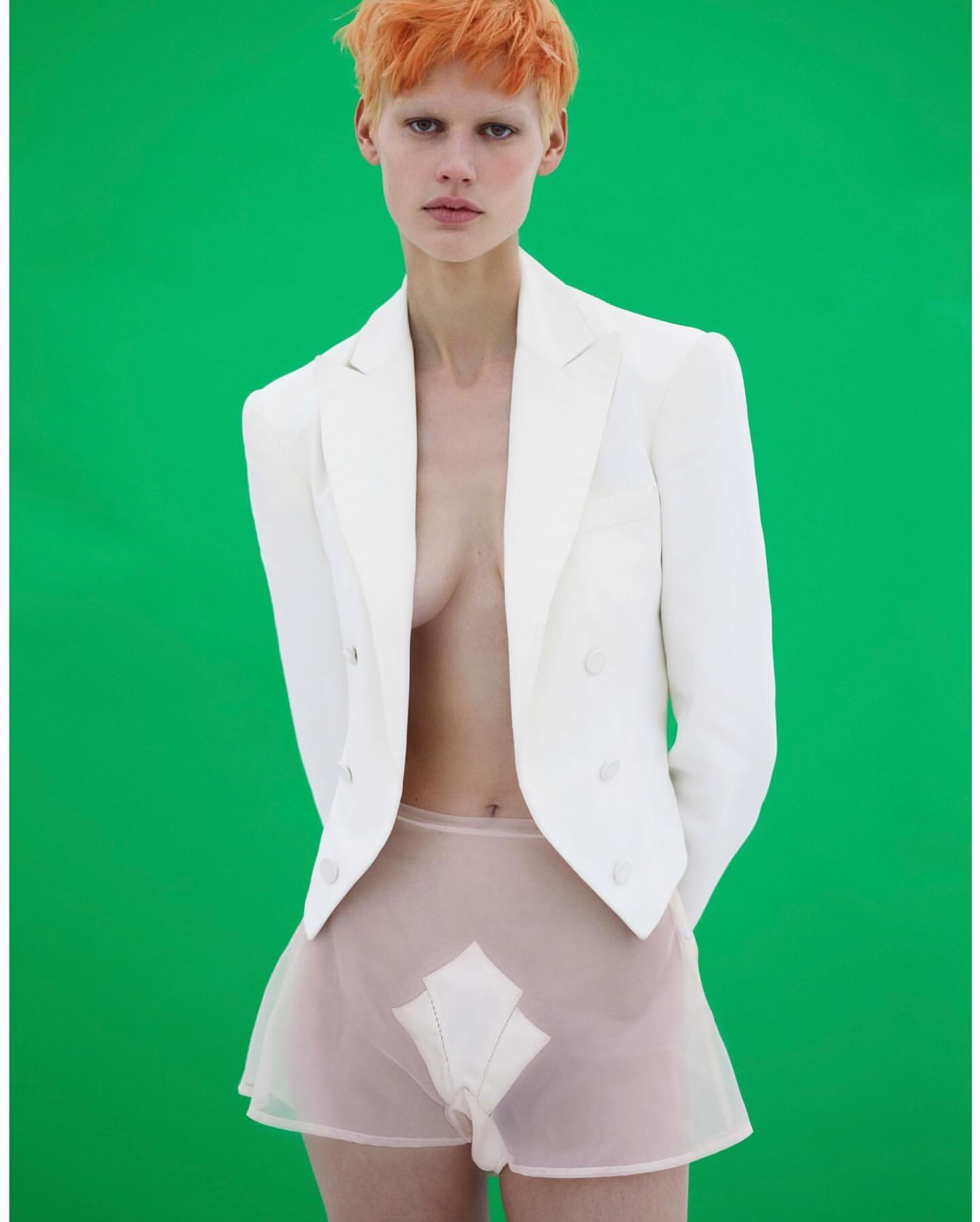 A pale-skinned woman with orange hair poses against a green backdrop, wearing a white blazer and sheer shorts in a studio setting.
