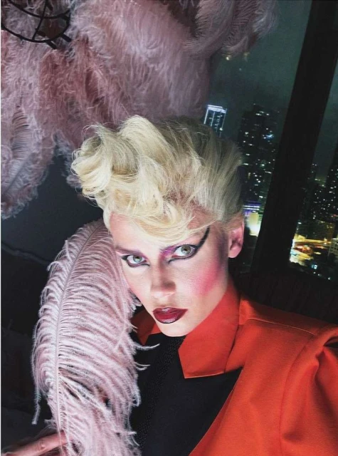 Woman with theatrical makeup and vintage hairstyle poses indoors with a feather fan and an orange jacket, with a city nightscape in the background.