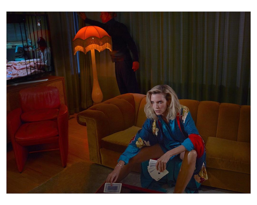 Blonde woman in robe plays cards on couch in an indoor living room scene with retro decor and a person standing in the background.