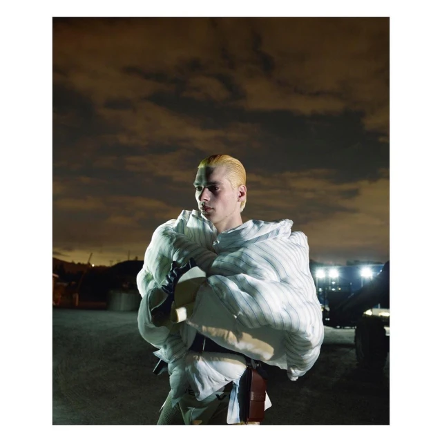 Indoor fashion shot of a blonde male model wearing a white pinstripe puffy jacket, with a cloudy, nighttime background and neutral tones.