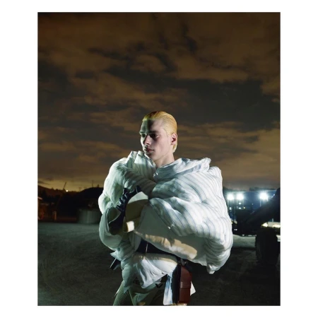 Indoor fashion shot of a blonde male model wearing a white pinstripe puffy jacket, with a cloudy, nighttime background and neutral tones.