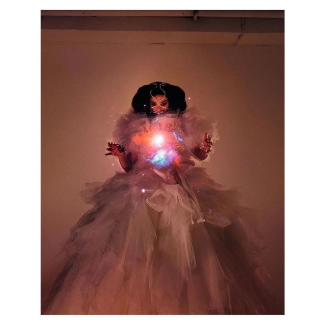 A woman in an elaborate tulle gown holds a glowing orb, standing in front of an orange background in an artful portrait.