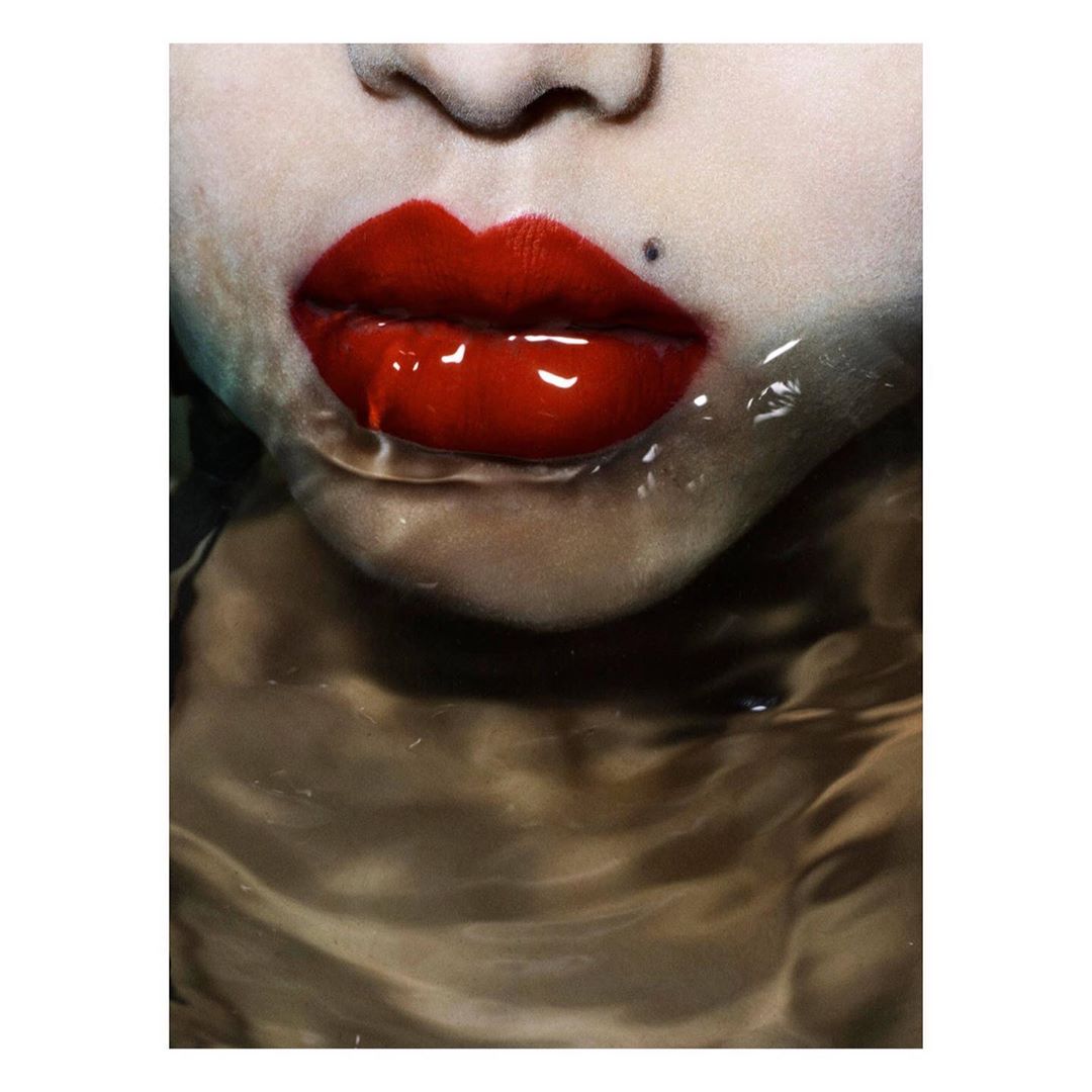 Close-up of a woman's face with glossy red lipstick, partially submerged in water creating reflections and shimmering effects.