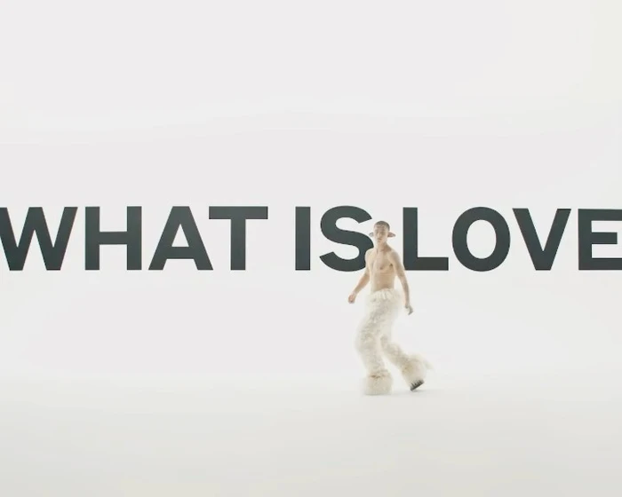 Shirtless person in furry white trousers walking past large bold text reading WHAT IS LOVE on white seamless background