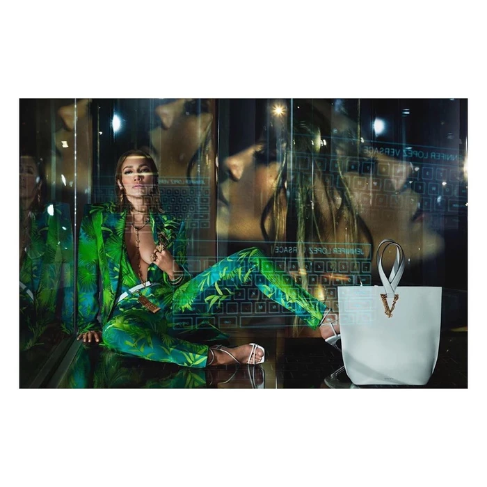 Jennifer Lopez seated wearing a green tropical print suit with a white handbag in foreground