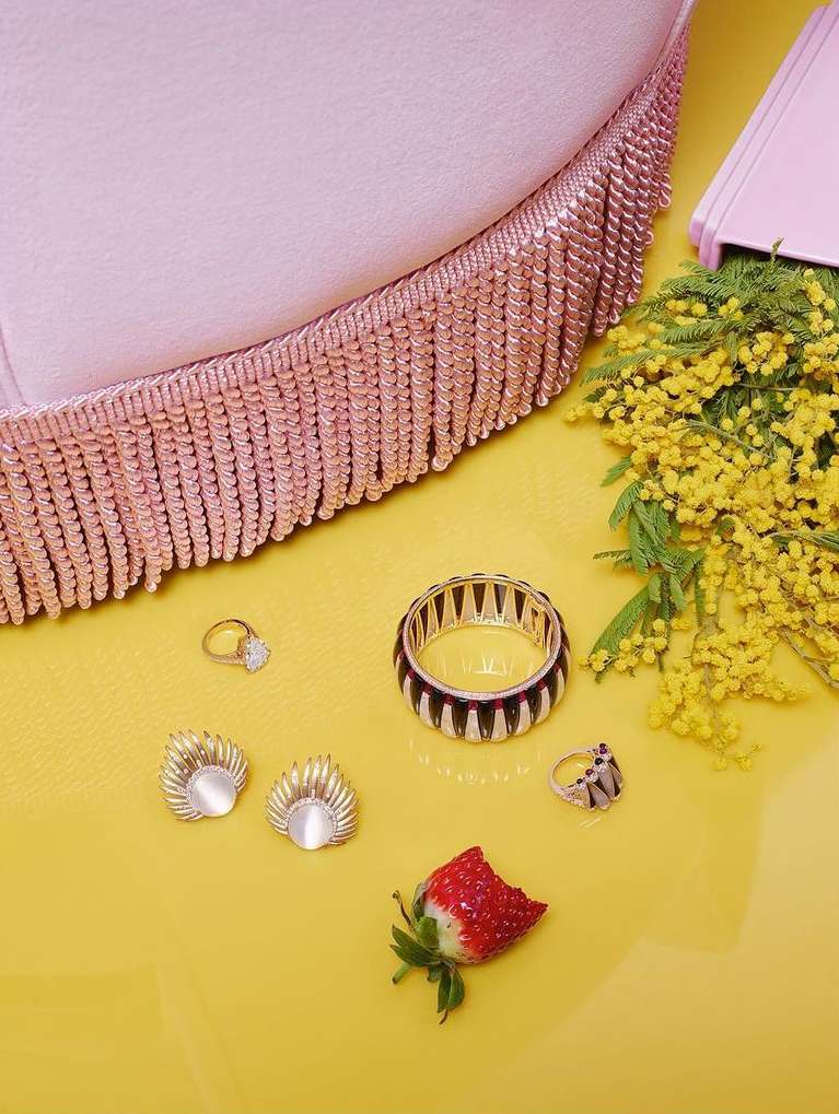 Still life of jewelry including rings and earrings with a strawberry and yellow flowers on a yellow surface next to a pink fringed object.