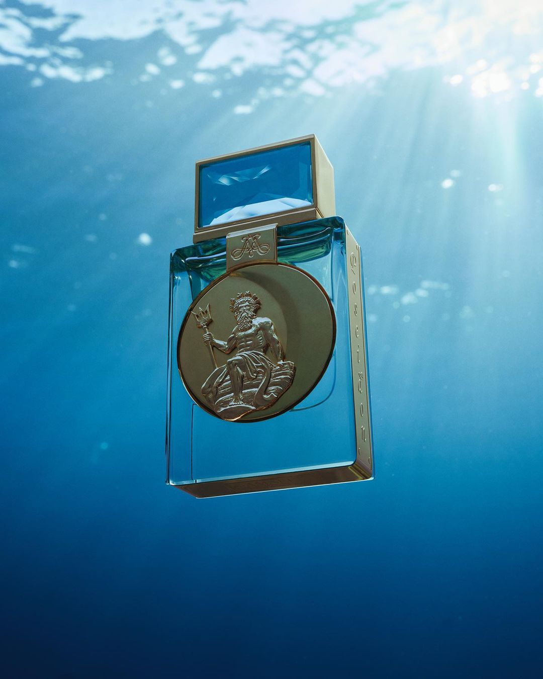 Elegant product shot of a clear blue glass perfume bottle with gold accents and embossed figure of Poseidon against a blurred blue underwater background.