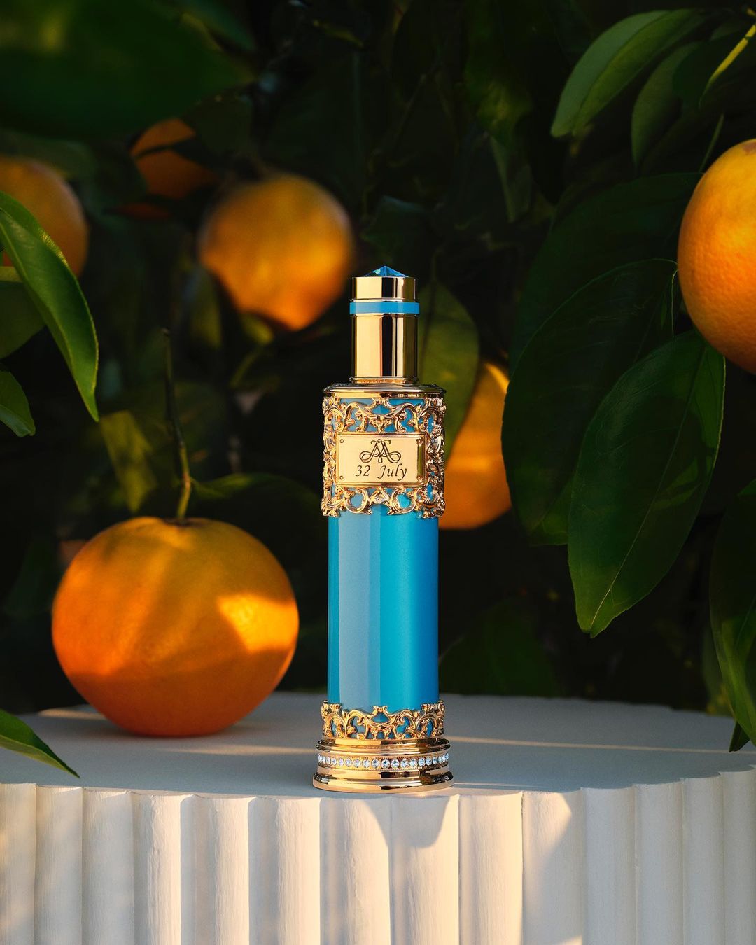 Luxurious turquoise and gold perfume bottle sits on a white fluted pedestal, surrounded by orange tree leaves and oranges in natural light.