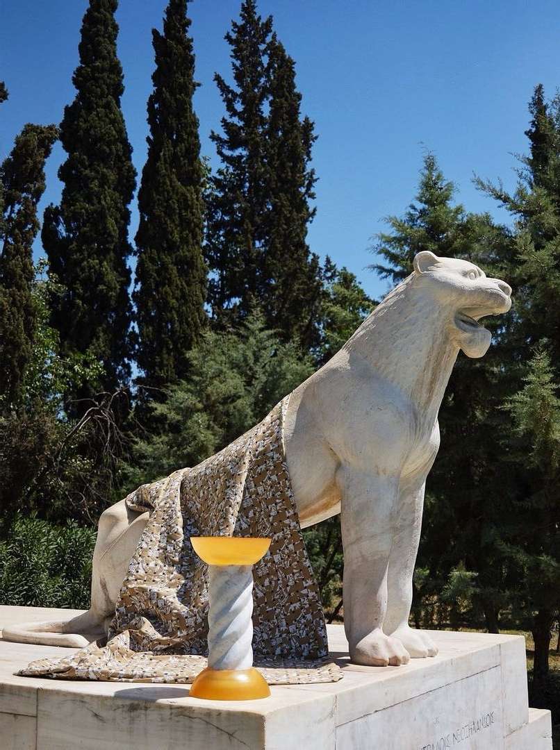 Marble sculpture of a roaring lion with a patterned drape, and an amber glass bowl on a pedestal in an outdoor park setting.
