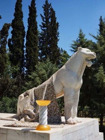 Marble sculpture of a roaring lion with a patterned drape, and an amber glass bowl on a pedestal in an outdoor park setting.