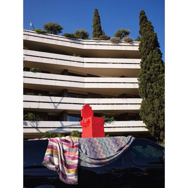 Modern building with stacked balconies, cypress trees, and a red sculpture. Blankets draped over a dark van in the foreground.