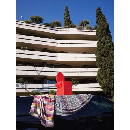 Modern building with stacked balconies, cypress trees, and a red sculpture. Blankets draped over a dark van in the foreground.
