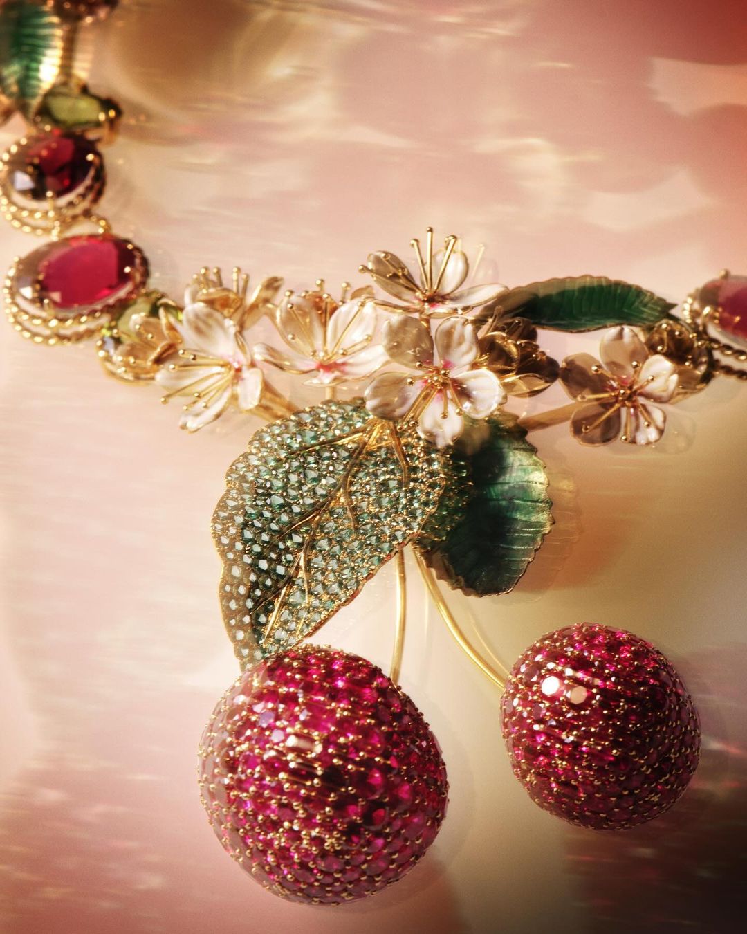 Close-up of an elaborate golden necklace featuring cherry and floral designs, adorned with pink gemstones and set against a soft backdrop.