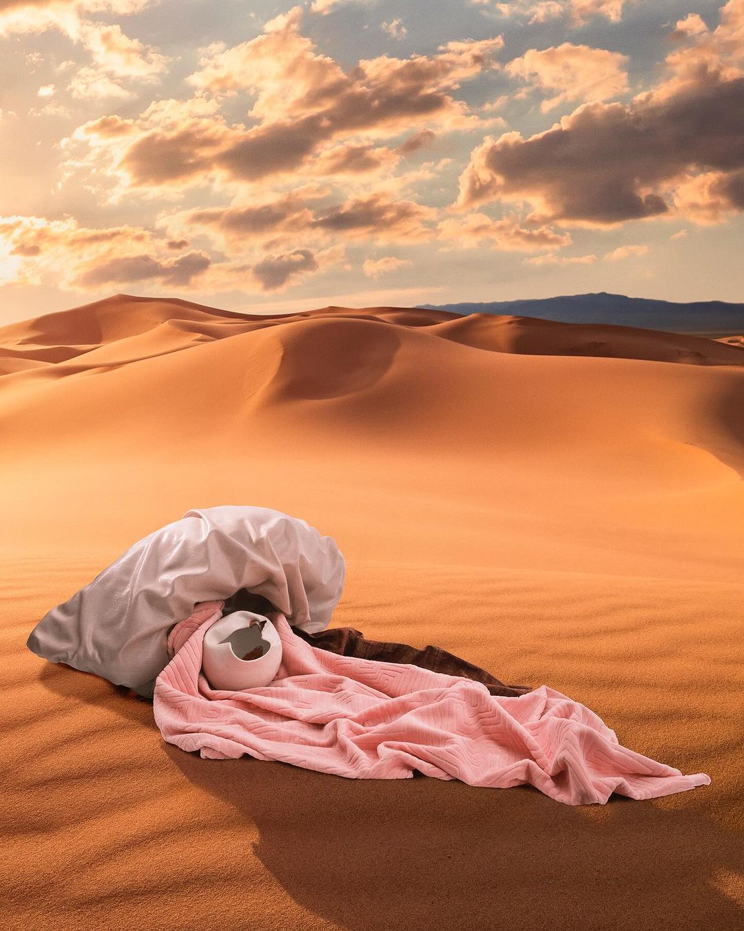 Desert landscape with draped bedding and a broken vase on sand dunes under a vibrant sky with golden-hour sunlight and scattered clouds.