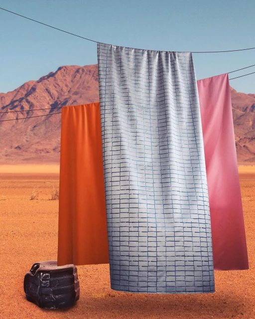 Three colorful fabric sheets hang from a wire in a desert landscape with mountains and blue sky in a surreal artistic composition.