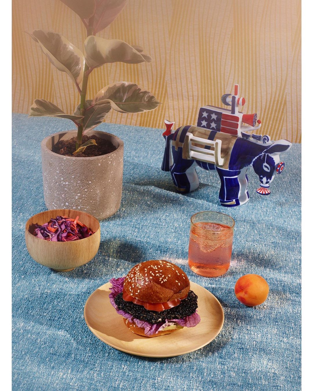 Still life with a burger topped with black garlic, a decorative donkey figurine, and a potted plant against a patterned background.