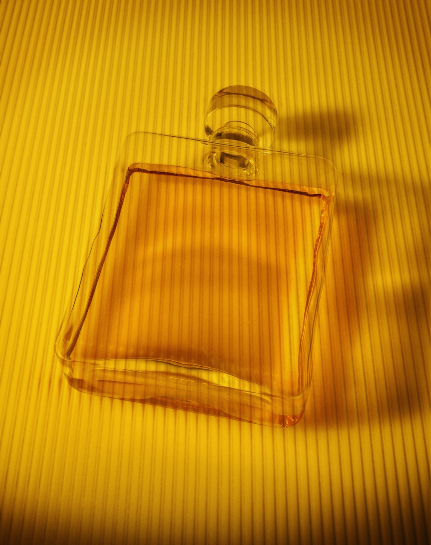 Transparent square perfume bottle filled with amber liquid, featuring a round stopper, set against a ridged gold background.