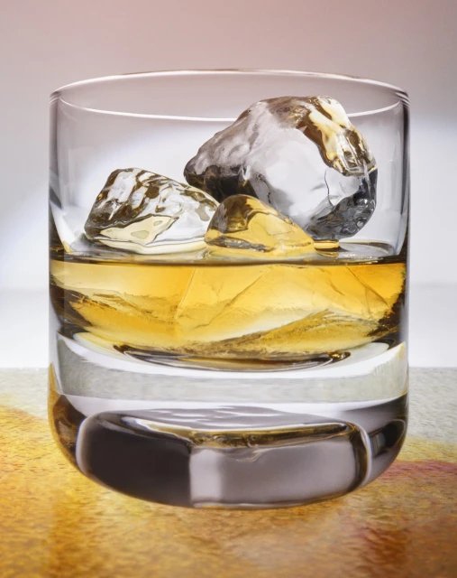 Close-up of whiskey in clear glass with large ice cubes against a blurred background, studio lit, emphasizing texture and color.