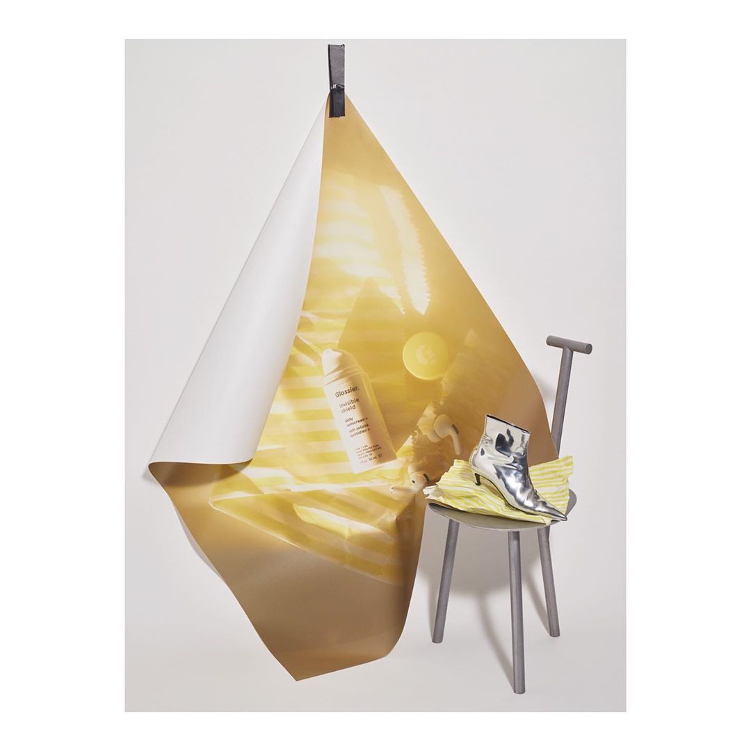 Still life composition featuring a beauty product, yellow fabric, and a silver ankle boot on a gray chair against a white backdrop.