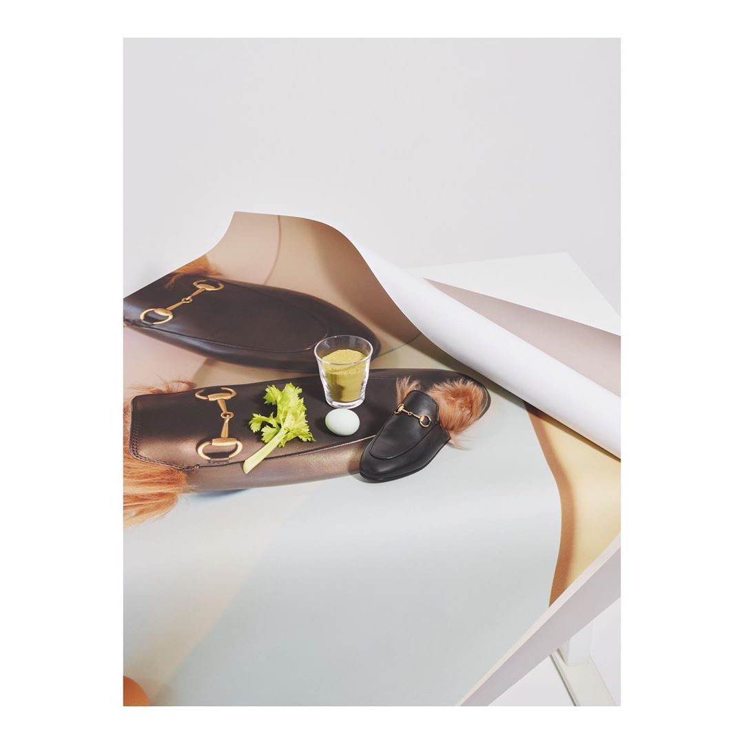 Still life composition of a designer loafer with fur trim, celery stalks, a drink, and a clutch on a white surface with a rolled backdrop.
