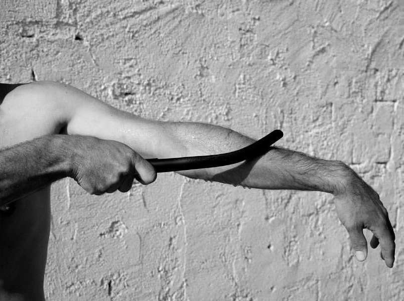 Monochromatic close-up of an adult man's arm holding a dark, curved stick in front of a textured white wall, focusing on skin and shadow.