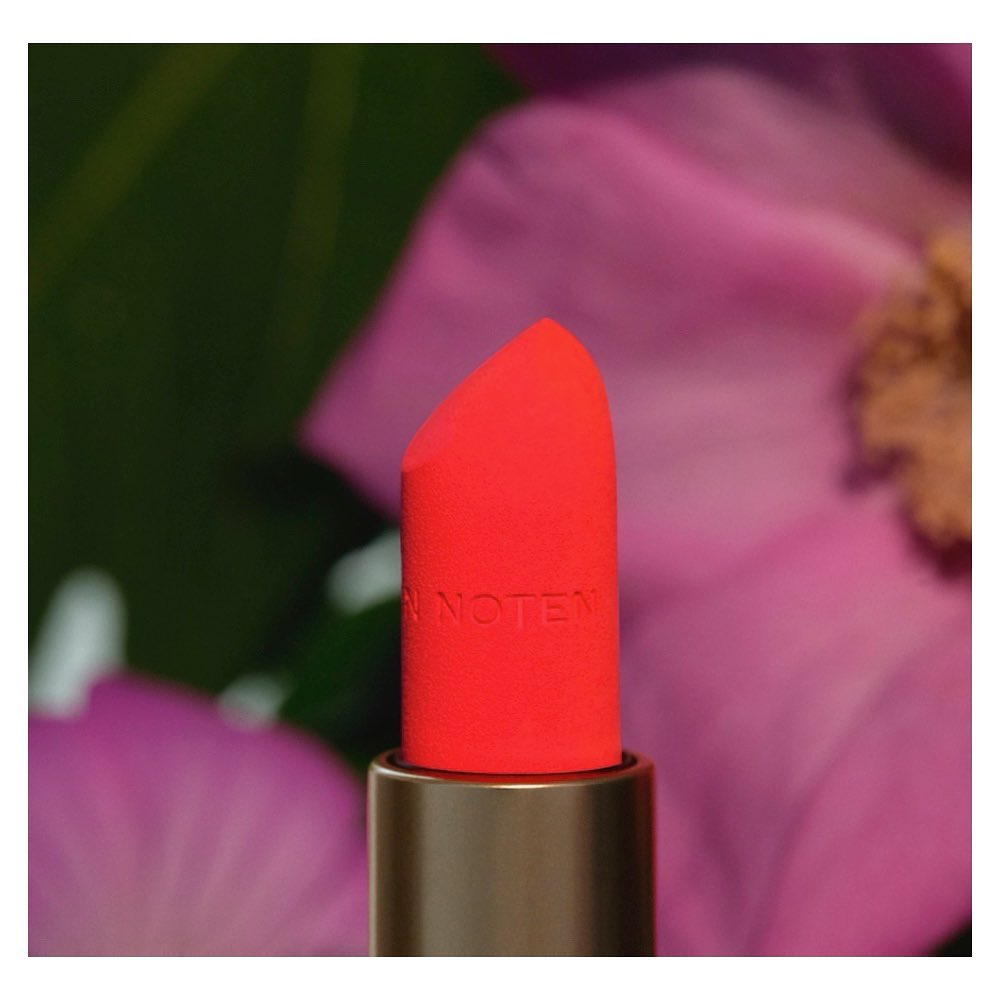 Macro shot of vibrant coral lipstick with gold tube against blurred floral background in soft focus for beauty and cosmetic marketing.