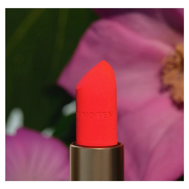 Macro shot of vibrant coral lipstick with gold tube against blurred floral background in soft focus for beauty and cosmetic marketing.