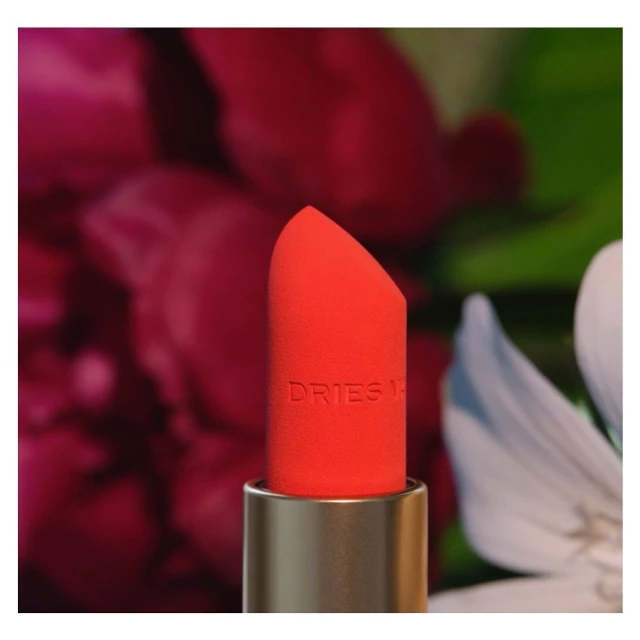 Close-up of coral lipstick tube with gold base featuring the brand name Dries Van Noten against a blurred floral background.