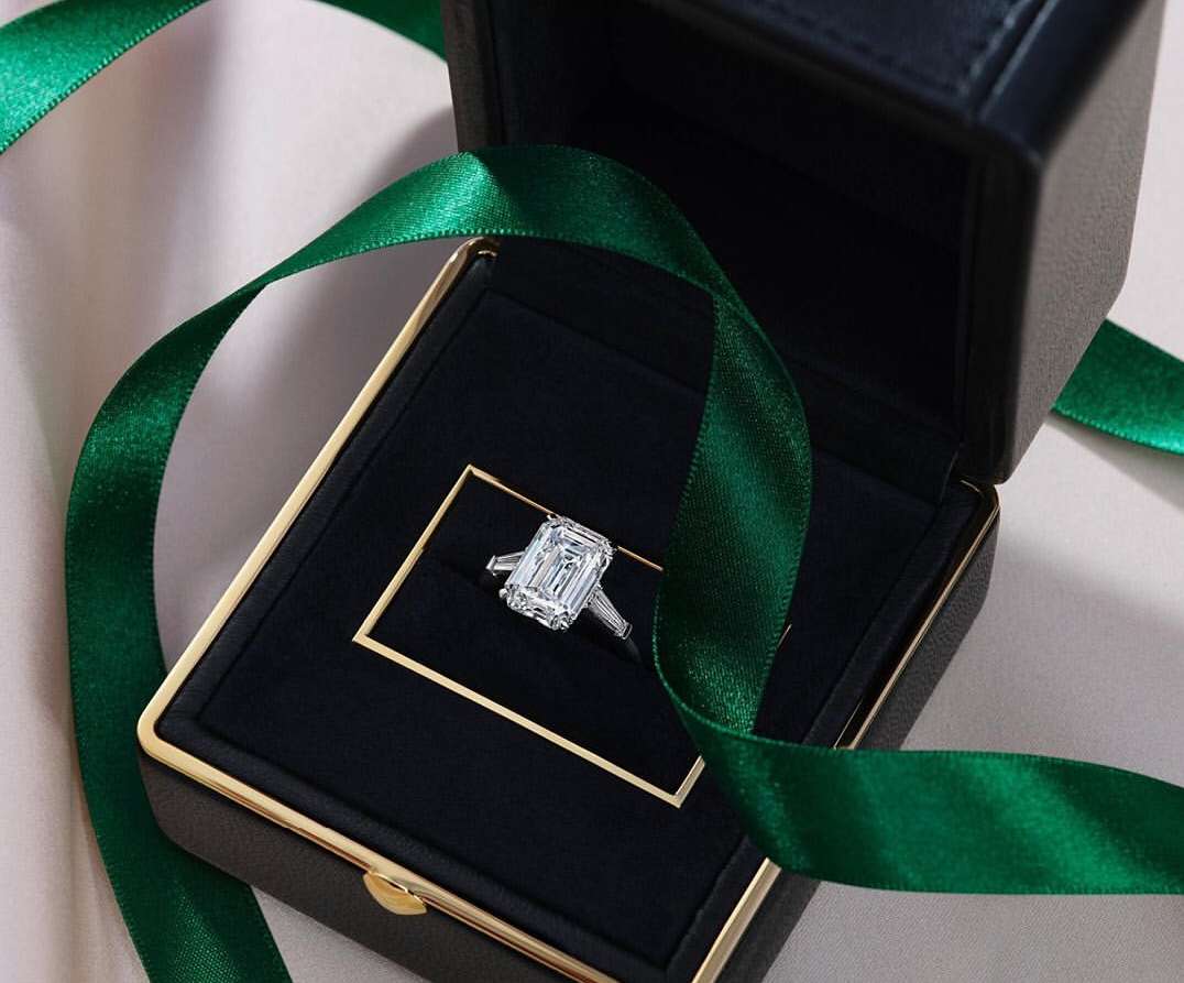Elegant emerald-cut diamond engagement ring in a black jewelry box with a gold frame, accented by a green ribbon on a light surface.