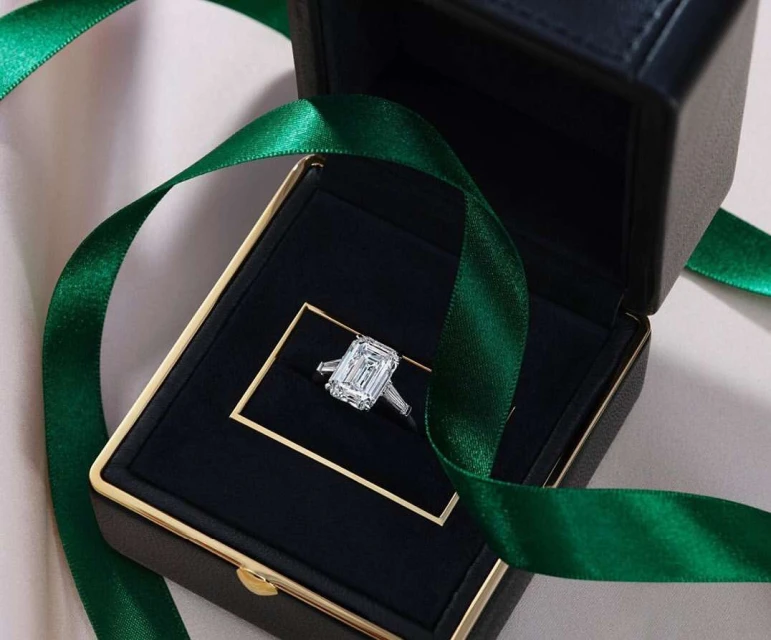 Elegant emerald-cut diamond engagement ring in a black jewelry box with a gold frame, accented by a green ribbon on a light surface.