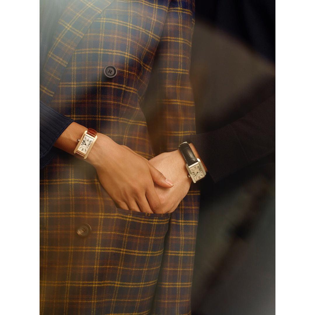 Close-up of two people holding hands, wearing wristwatches and suit jackets with a plaid pattern, against a dark background.