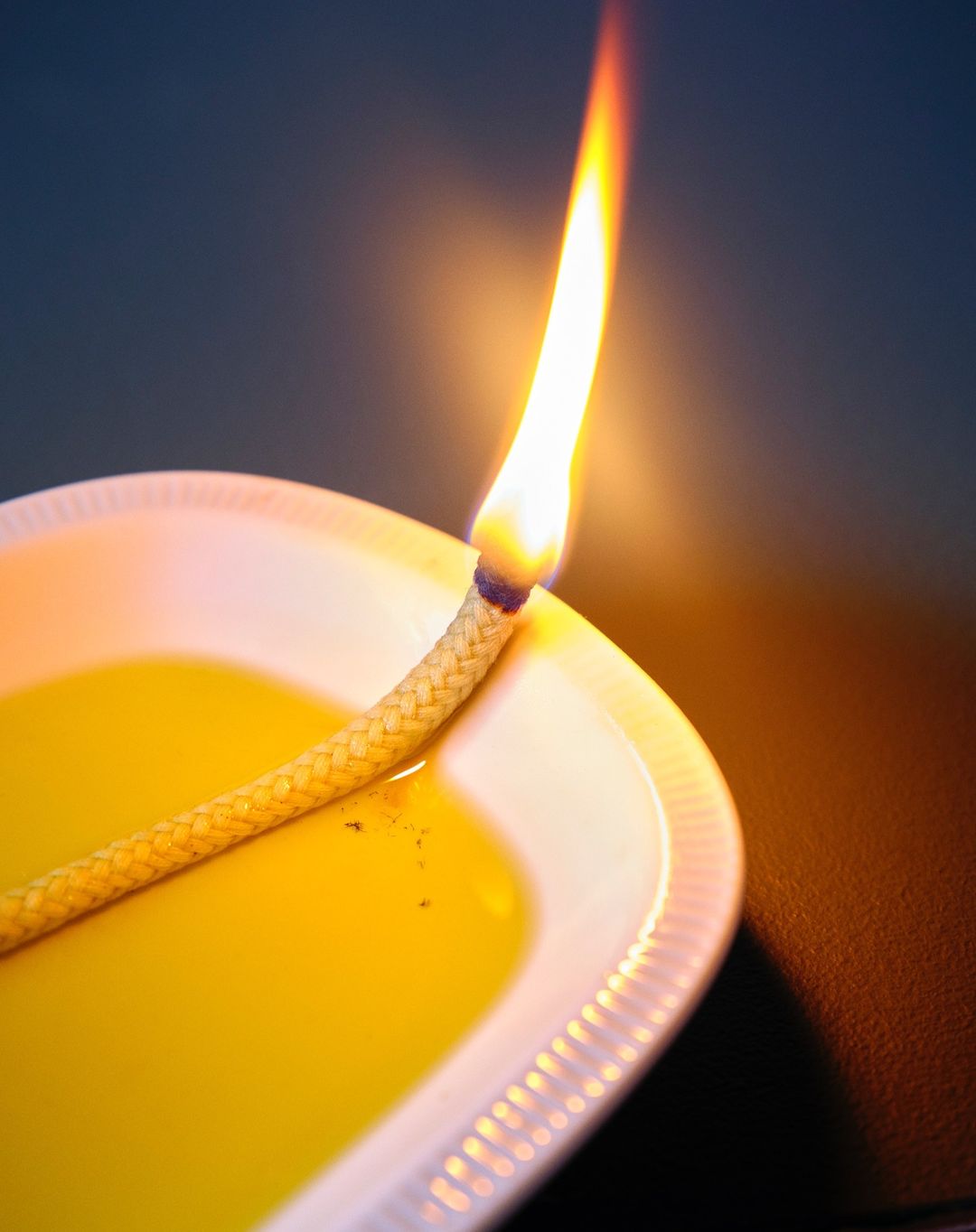 Close-up of a lit diya lamp with a bright flame, featuring a cotton wick in oil on a white plate against a dark background.