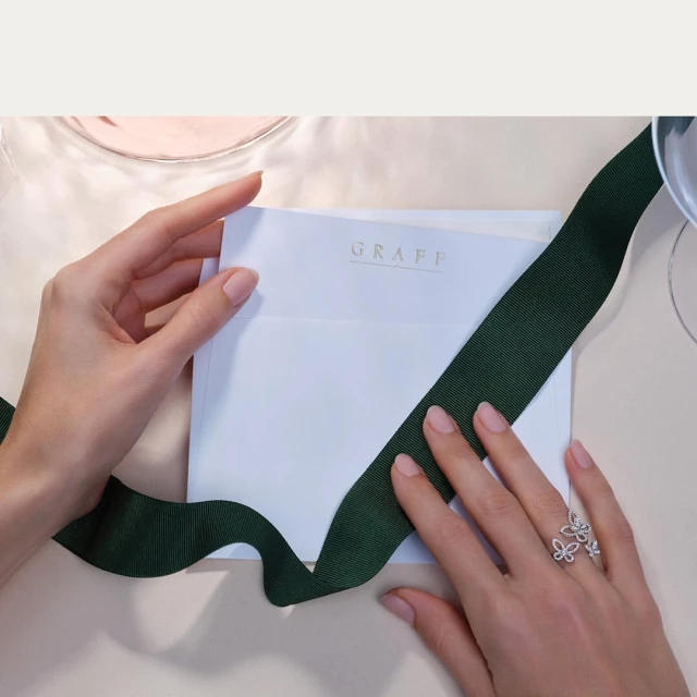 Close-up of manicured hands holding a white Graff card with a green ribbon and floral diamond ring, set on a neutral surface.